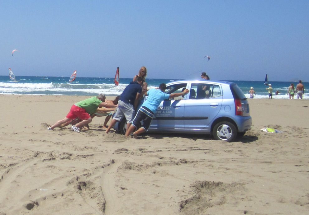 People trying to push a car stuck in the sand on Prasonissi Beach. Click for Travel Insurance